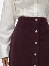 Saia de corduroy bordeaux com cintura alta, apresentando cinco botões de metal na parte da frente e uma textura suave. Camisa branca de mangas longas em cima.