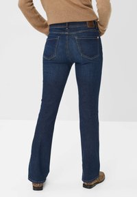 Jeans flared in denim blu scuro con vita alta, tasche posteriori e cuciture a contrasto. Tessuto flessibile e leggera texture visibile.