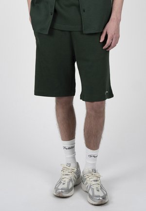 Mann trägt dunkelgrüne Shorts, weiße Sportsocken mit schwarzem Text und weiße und graue Sportschuhe, steht auf einem einfarbigen Hintergrund.