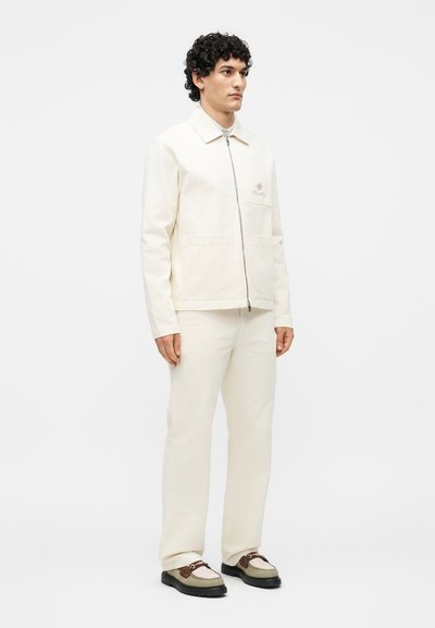 Άνδρας με σγουρά μαλλιά που φοράει μπεζ-λευκό μπουφάν με φερμουάρ, ταιριαστό παντελόνι και μπεζ-καφέ μοκασίνια, στέκεται μπροστά σε απλό φόντο.