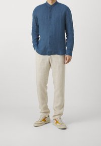 Hombre con una camisa de botones azul de manga larga, pantalones de lino beige y zapatillas beige con acentos en naranja y amarillo, de pie contra un fondo liso.