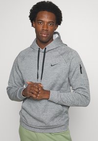 Grå hættetrøje med struktureret stof, snor i huen og en lynlåslomme på ærmet; har et sort Nike-logo på brystet.