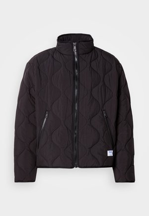 Veste noire matelassée avec un motif de coutures ondulées, fermeture éclair à l'avant, deux poches latérales zippées, et une petite étiquette de marque blanche près de l'ourlet.