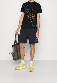 Muž v černém tričku Karl Lagerfeld s vícebarevným potiskem, černých šortkách Dondup, bílých ponožkách, žlutých teniskách a držící zelenou a modrou tašku.