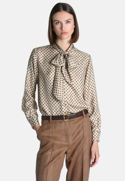 Femme portant un chemisier beige à motifs avec un grand nœud papillon et un pantalon marron taille haute avec une ceinture foncée, debout avec une main dans la poche.