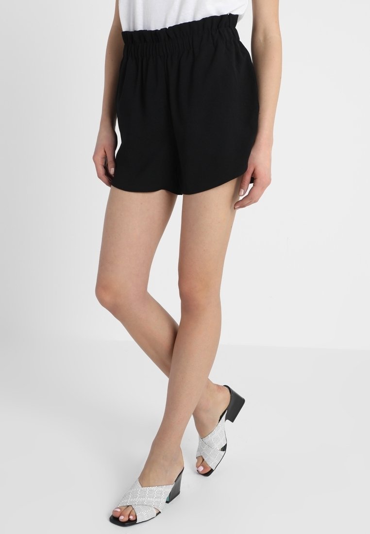 Femme portant un short noir à taille élastique et des sandales blanches à talons avec des lanières croisées, debout jambes croisées sur fond blanc.
