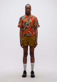 Γυναίκα που στέκεται και φοράει πορτοκαλί κοντομάνικο πουκάμισο με λουλούδια, χακί cargo σορτς, λευκές κάλτσες και μαύρα αθλητικά παπούτσια σε λευκό φόντο.