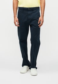 Marineblå bomullsbukser med rett passform og frontlommer. Har knappelukking og beltesløyfer, kombinert med hvite joggesko.