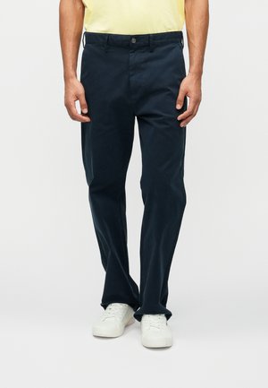 Mørkeblå bomuldsbukser med lige pasform og forlommer. Har en knaplukning og bæltestropper, kombineret med hvide sneakers.