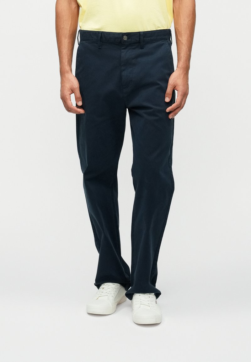 Marineblå bomullsbukser med rett passform og frontlommer. Har knappelukking og beltesløyfer, kombinert med hvite joggesko.