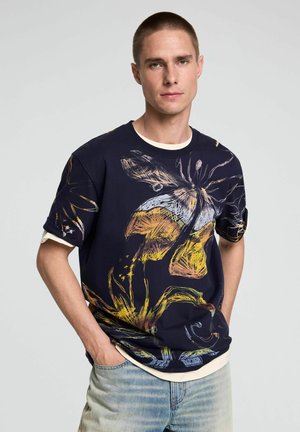 Mann mit kurzem Haar, der ein dunkelblaues T-Shirt mit großem farbenfrohem abstraktem Blumen- und Schmetterlingsmuster trägt, helle Jeans, Hände in den Taschen, neutraler Hintergrund.