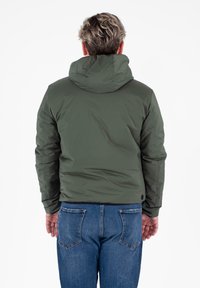 Veste rembourrée vert olive avec capuche, offrant une texture lisse et une coupe légèrement ajustée. Portée avec un jean en denim bleu.