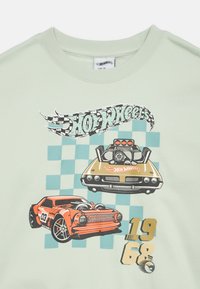 Maglietta verde chiaro con grafiche Hot Wheels che mostra due auto. Una è arancione, l'altra è dorata, con uno sfondo a scacchi e l'anno "1968."