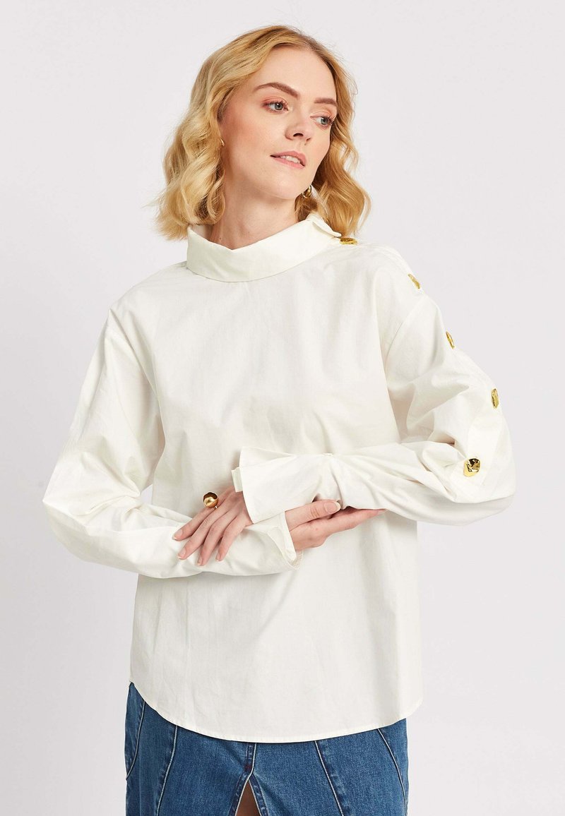 Kuldsete nööpidega valget pikkade varrukatega pluusi kandnud blond juukseline naine, seljas sinine teksaseelik, millel on ees lõhik.