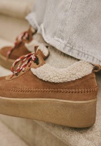 Brown suede platform shoes with faux fur collar, red and white laces, and decorative stitching. Textured rubber sole visible.