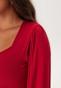 Röd topp med djup V-ringning, långa ärmar med draperad detalj vid axlarna och ett mjukt, stretchigt tyg.