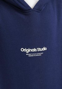 Tamno plava dukserica od mekog materijala, s bijelim logotipom "Originals Studio" i dodatnim oznakama branda blizu izreza.