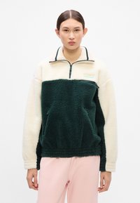 Fleecasti pulover s pola zatvaračem, zeleno tijelo, kremaste rukave, prednji džep i ovratnik na pruge. Ima malu oznaku ilišće na grudima.