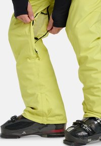 Handen die gele skibroek openen om mesh ventilatie over zwarte skischoenen met gespen te onthullen, op een witte achtergrond.