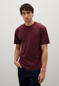 Joven con cabello castaño ondulado, vistiendo una camiseta de bolsillo burdeos sencilla y jeans azul oscuro, de pie contra un fondo neutro claro.