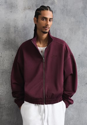 Mand med fletninger iført en vinrød lynlåstrøje og hvide bukser, stående foran en tekstureret grå væg.