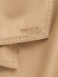 Un tissu beige avec une texture lisse présente une broderie "SL" et un détail de nœud. La couture est soignée, mettant en valeur un savoir-faire de qualité.