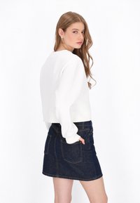 Fehér, rövidített pulóver hosszú ujjakkal, sötét farmer mini szoknyával kiegészítve, melynek hátsó zsebei és látható varrási részletei vannak.