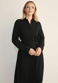 Kobieta z blond włosami, ubrana w czarną sukienkę z długim rękawem i zapięciem na guziki, z paskiem w talii, stojąca na jednolitym jasnym tle.