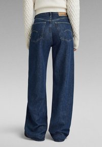 Jeans din denim închis la culoare, cu picioare largi și talie înaltă, având două buzunare la spate și un eticheta cu marcă la talie. Textura este netedă.