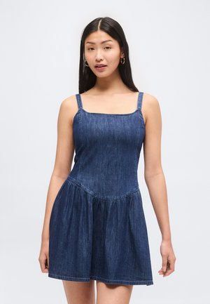 Kvinde med langt mørkt hår iført ærmeløs blå denimkjole med firkantet udskæring og samlet nederdel, stående mod en ensfarvet baggrund.