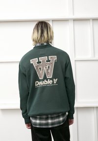 Grøn sweatshirt med et stort hvidt og brunt stribet logo "W" og "Double A" på ryggen; har en ribbet krave og struktureret stof.