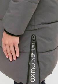 Grå pufferjakke med en glatt tekstur. Har en kontrastfarge svart glidelås, logodetaljer og en sidelomme.