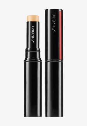 Bâton de maquillage noir avec un noyau beige clair, de forme cylindrique, finition brillante, portant le logo Shiseido et une ligne d'accent rouge sur un côté.