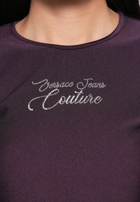 Detailní záběr na osobu, ktorá má na sebe tmavofialový top s nápisom "Versace Jeans Couture" vyšitým kamienkami na hrudi.