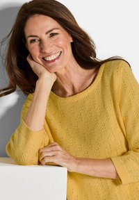 Gelber Pullover mit lockerer Passform, schaufelähnlichem Ausschnitt und strukturiertem Muster. Modell mit der Hand auf einer weißen Oberfläche ruhend, zeigt das entspannte Design.