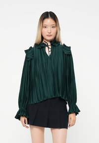 Blusa plissettata verde smeraldo con collo alto, dettagli a volant sulle spalle e maniche lunghe, abbinata a una gonna plissettata nera.