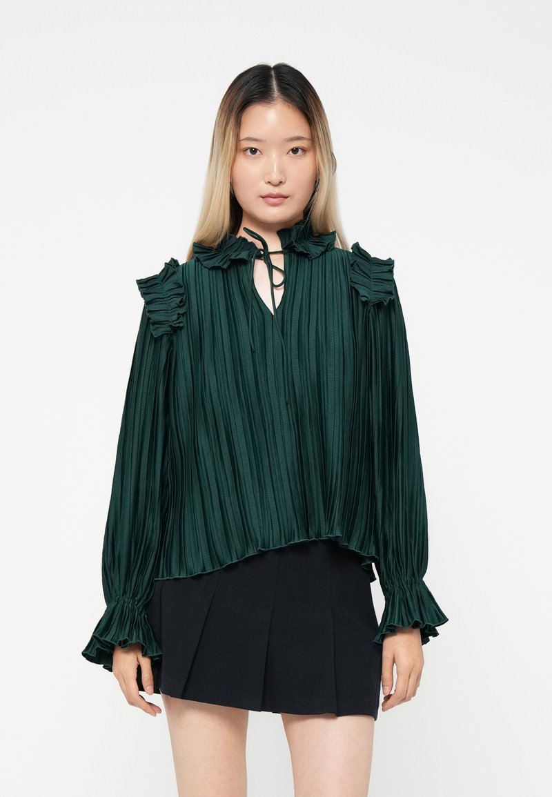 Blusa plissettata verde smeraldo con collo alto, dettagli a volant sulle spalle e maniche lunghe, abbinata a una gonna plissettata nera.