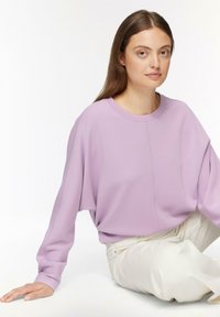 Sudadera oversized de color lavanda con cuello redondo, hombros caídos y detalles de costura sutiles, combinada con pantalones de color crema.