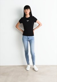 Camisa negra ajustada con un pequeño logo blanco en el pecho, combinada con jeans skinny de un azul claro y zapatillas blancas, frente a un fondo blanco.