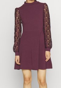 Burgundy klänning med hög krage, figursydd överdel och långa genomskinliga spetsärmar med ett diamantmönster. Mjuk textur, längd över knä.