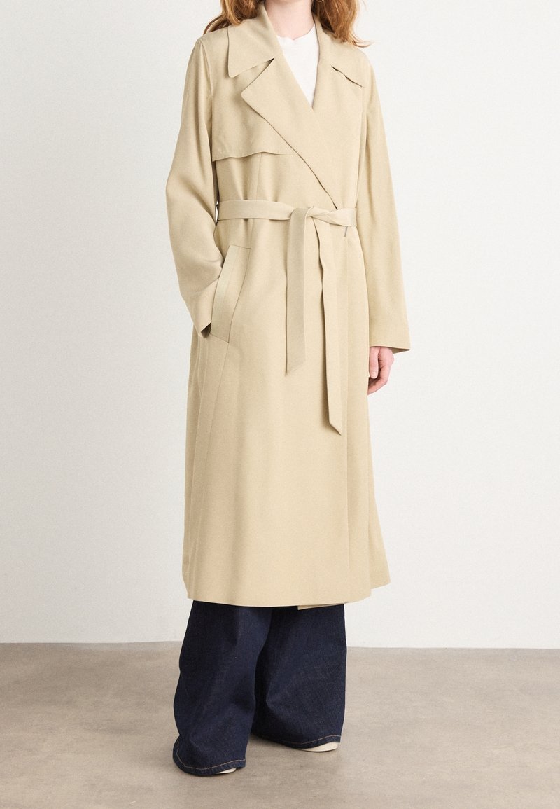 Personne portant un trench-coat beige ceinturé avec de larges revers sur un pantalon large sombre, debout sur un sol neutre devant un mur uni.