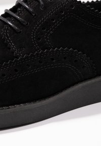 Scarpe brogue in suede nero con intricate perforazioni, un motivo ondulato lungo il bordo superiore e una suola piatta nera.