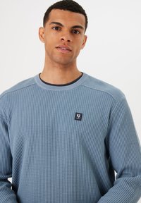 Lichtblauwe gebreide trui met een patroon, ronde halslijn en een klein vierkant logo op de borst. Lange mouwen met geribbelde manchetten.