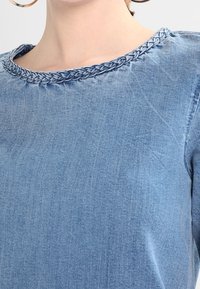 Vaaleansininen farkkutoppi, jossa on letitetty pääntie. Silkkinen pinta, yksinkertainen muotoilu ja lyhyet hihat.