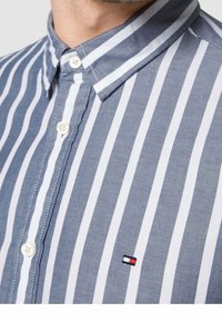 Primo piano di una camicia a righe verticali blu e bianche con un piccolo logo Tommy Hilfiger sul petto.