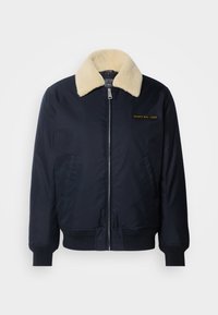 Chaqueta bomber navy con un cuello de piel sintética color crema, bolsillos laterales, puños de canalé, cierre frontal con cremallera y logo bordado en dorado en el pecho.