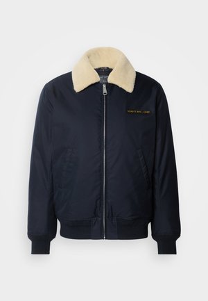 Chaqueta bomber navy con un cuello de piel sintética color crema, bolsillos laterales, puños de canalé, cierre frontal con cremallera y logo bordado en dorado en el pecho.