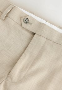 Gros plan de la taille d’un pantalon beige avec fermeture par bouton et passant de ceinture, montrant la texture du tissu et les détails de couture.