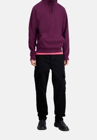 Sweat à capuche bordeaux avec une poche avant et une capuche à cordon, porté sur un t-shirt rose. Associé à un pantalon cargo noir avec des poches latérales. Chaussures noires.