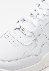 Vit lädersneaker med perforeringar på tån, vävda skosnören och en metallisk logotypetikett. Slät textur med kontrasterande sömmar.